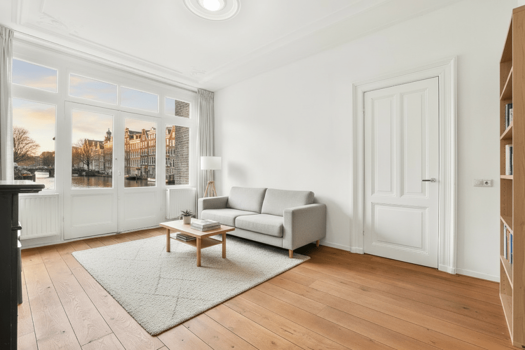 Take this exact empty Amsterdam apartment room with white walls, white ornate ceiling, large French windows with white curtains, warm oak hardwood floors, and white paneled door. Furnish it with Scandinavian-modern style furniture: a light grey linen sofa, a wooden coffee table with books and a small plant, a floor lamp, a minimal bookshelf against the right wall, and a soft area rug. Through the large French windows, replace the current brick building view with a stunning panoramic view of Amsterdam canals at golden hour — with canal houses, water reflections, and warm sunlight. Keep the room architecture identical, only add furniture and change the window view.